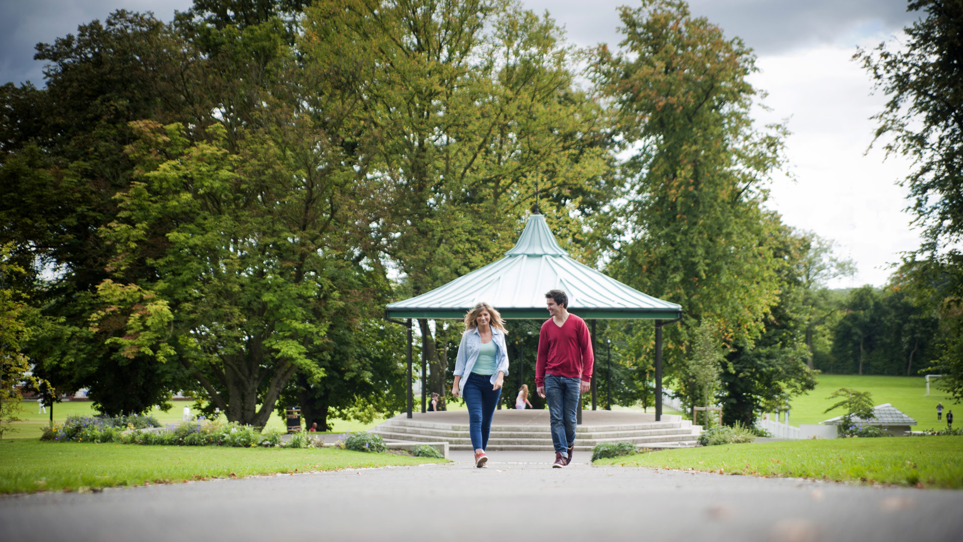 A wide, open green expanse of Wallace Park in Lisburn, with pathways, mature trees, and a distant pavilion under a bright sky.